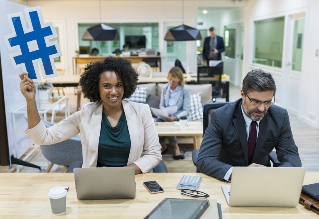 In an office, woman holds up a sign that looks like a hashtag, a useful tactic in brand activation